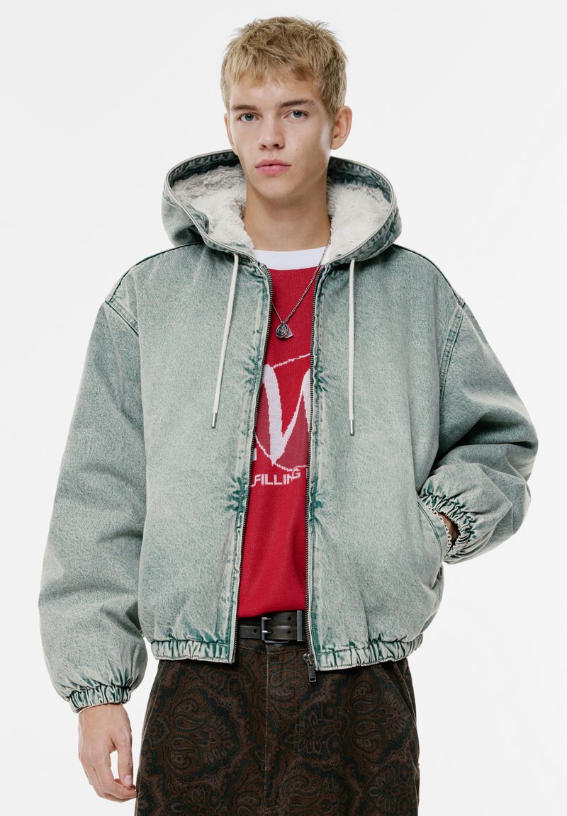 Geacă din denim verde cu un guler căptușit cu blană, manșete elastice și brâu elastic. Purată deasupra unei tricouri grafic roșii și a unor pantaloni cu model.