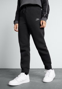 Svarta joggerbyxor med en mjuk textur, med resår i midjan och i bensluten, samt en liten grå Nike-logga på vänster lår. Bärs med vita sneakers.