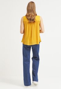 Top giallo senza maniche con schiena arricciata, abbinato a jeans dritti blu scuro e sneakers bianche. Design semplice e stile casual.