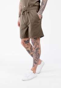 Homme portant un short à cordon brun avec des tatouages sur les jambes et les bras, associé à des baskets blanches, se tenant devant un fond blanc.