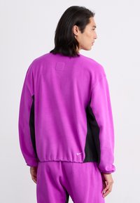 Violet spalvos fliso džemperis su juodomis šoninėmis juostomis, raštuotais rankogaliais ir išsiuvinėtu logotipu apatine dalyje. Lygi tekstūra ir atsipalaidavęs kirpimas.