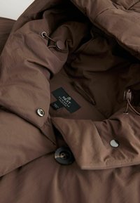 Bruine outerwear met een gladde textuur, voorzien van een knoopsluiting en een verstelbaar trekkoord. Label zichtbaar aan de binnenkant met merkinformatie.
