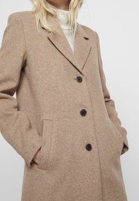 Donna che indossa un cappotto di lana beige con tre bottoni neri e mani nelle tasche laterali sopra un maglione a collo alto bianco.