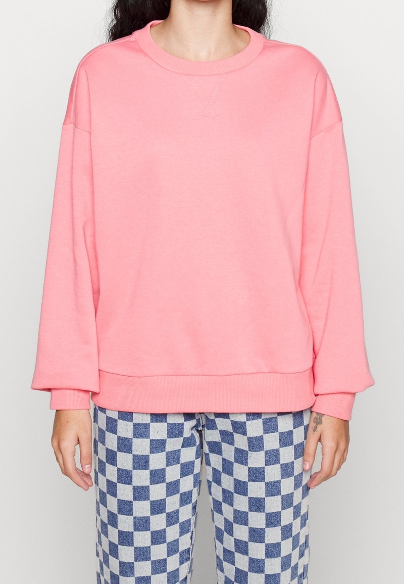 Femme portant un sweat-shirt rose ample et un pantalon à carreaux bleus et blancs, debout devant un fond clair uni.