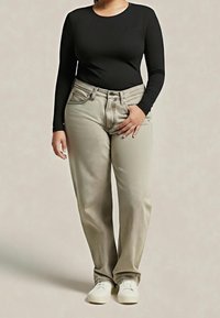 Personne portant un haut noir à manches longues, un pantalon beige clair et des chaussures blanches, debout avec une main dans la poche sur un fond neutre.