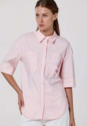 Femme portant une chemise ample rose pâle à boutons avec des manches jusqu'au coude et un petit motif floral sur la poche poitrine gauche.