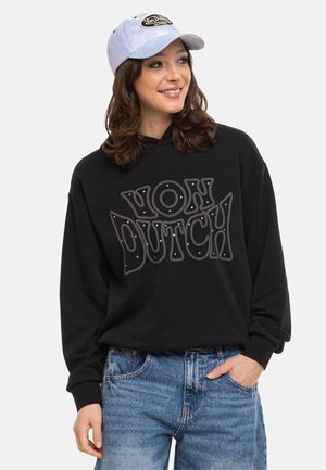 Femme portant un sweat à capuche noir "Von Dutch", une casquette de baseball bleu clair et un jean bleu, souriante et regardant légèrement vers la droite.