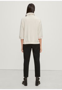 Lichtbeige pullover met een hoge kraag en een oversized pasvorm, voorzien van driekwart mouwen en een gerimpelde tailleband, gecombineerd met zwarte broeken.