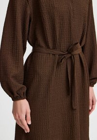 Robe marron texturée avec des manches longues, des poignets élastiques et une ceinture à la taille pour un ajustement réglable. Le tissu présente un effet légèrement froissé.