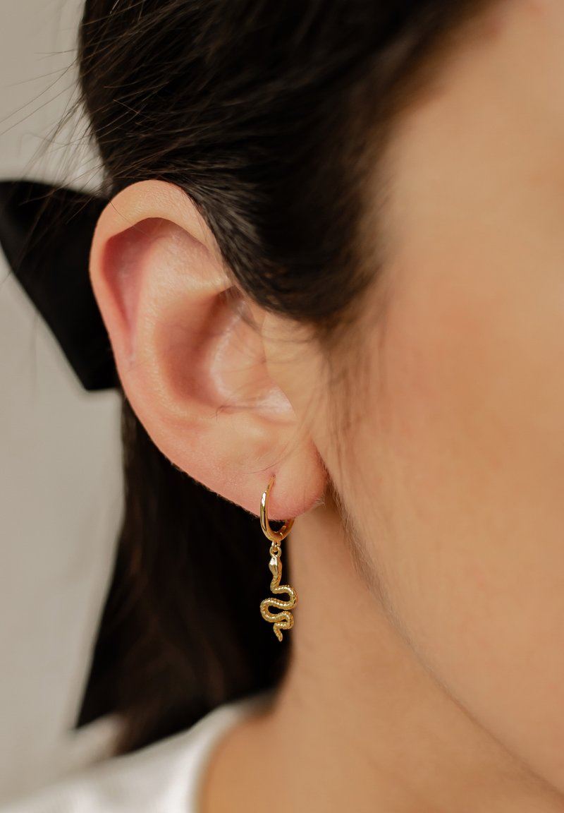 Pendiente de aro de oro con diseño de serpiente, con un detallado intrincado y un acabado pulido, colgando de la lóbulo de la oreja sobre un fondo claro.