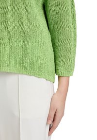 Hellgrüner, gestrickter Pullover mit strukturiertem Muster, Dreiviertelärmel und einem leicht verkürzten Saum, getragen über weißen Hosen.