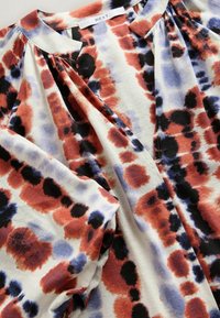 Blouse avec encolure froncée et taches abstraites multicolores en rouille, noir et lavande sur fond blanc.