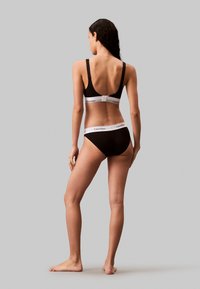 Soutien-gorge en coton noir et culotte assortie avec des bandes blanches portant le logo Calvin Klein. Le soutien-gorge dispose de bretelles réglables et d'une fermeture à crochet et œillet.