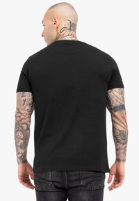 Schwarzes T-Shirt mit kurzen Ärmeln, aus Baumwollmaterial, figurbetontes Design, glatte Textur, keine sichtbaren Logos, kombiniert mit dunklen Jeans, Tattoos an den Armen.