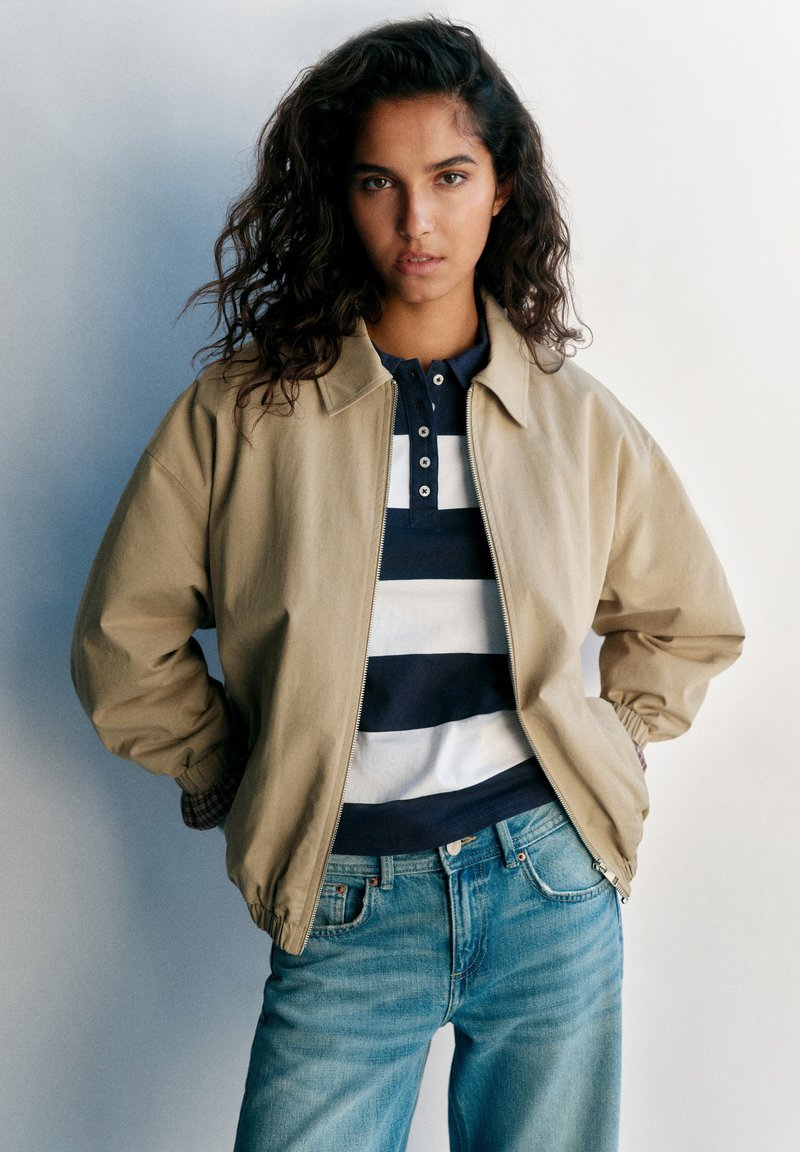 Jeune femme aux cheveux bouclés portant une veste beige, une chemise rayée bleu marine et blanche, et un jean bleu, debout les mains dans les poches devant un fond uni.
