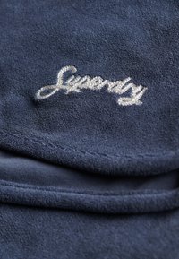 Tissu texturé bleu foncé avec le mot en cursive blanche brodé "Legendary" près du centre, plié avec des coutures et des bords visibles.