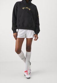 Sweat à capuche noir avec le texte jaune « PARIS », short blanc et chaussettes blanches montantes jusqu'aux genoux. Porté avec des crampons de football blancs et roses.