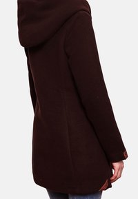 Cappotto marrone con cappuccio, dalla texture morbida, con il retro più lungo e dettagli con logo sui polsini. Presenta un design semplice e aerodinamico.