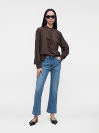 Femme portant un chemisier marron foncé à manches longues, un jean bleu droit, des chaussures noires pointues et des lunettes de soleil noires, debout avec une main dans la poche.