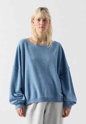 Vrouw draagt een losse, gestructureerde lichtblauwe trui met afgezette schouders en lichtgrijze broek, staand tegen een effen witte achtergrond.