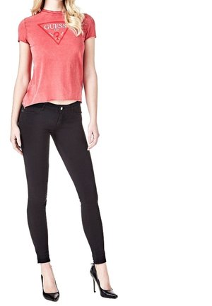 Kobieta w czerwonym T-shircie z krótkim rękawem i logo Guess, czarnych obcisłych jeansach oraz czarnych butach na wysokim obcasie, stojąca na białym tle.