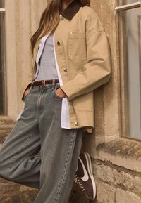 Beige kanavapusero, jossa on ruskea corduroy-kaulus, nappikiinnitys ja etutasku, yhdistettynä löysiin harmaisiin farkkuihin ja ruskeisiin/valkoisiin tennareihin.