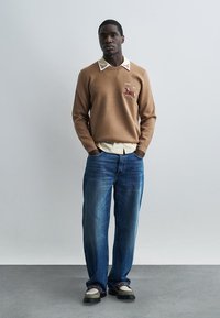 Hombre con suéter marrón con ciervo bordado, camisa crema con cuello, vaqueros azules y zapatos bicolor, de pie contra una pared lisa y clara.