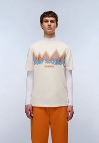 Kremfarvet t-shirt med bjerggrafik i orange og blå, båret over en hvid langærmet top. Kombineret med orange bukser. Simpelt design.