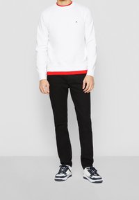 Hombre vistiendo un sudadera blanca con detalles rojos, camiseta interior roja, pantalones negros y zapatillas blancas con detalles en azul marino, de pie ante un fondo liso.