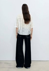 Transparent vit topp med blommig broderi, breda svarta denimbyxor med lös passform och en lätt slitna fåll.