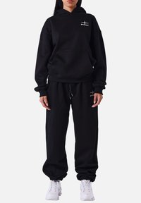 Conjunto de sudadera con capucha y pantalones de chándal, hecho de tejido suave. La sudadera cuenta con un bolsillo frontal y detalle del logo; los pantalones tienen puños elásticos y cordón ajustable.