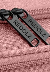 Tessuto rosa testurizzato con due tirazip neri etichettati "REDOLZ". Denti della zip visibili, che evidenziano il dettaglio della chiusura.