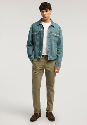 Joven de pie vistiendo una chaqueta vaquera azul, camiseta blanca, pantalones beige, cinturón marrón y mocasines marrones sobre un fondo liso.