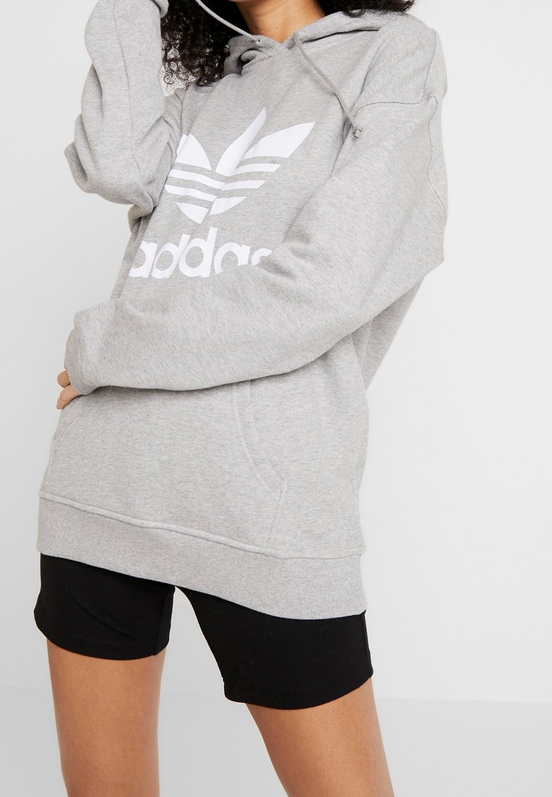 Grå huvtröja med vit Adidas-logotyp, tillverkad av mjukt tyg. Har en framficka och dragsko i huvan. Kombinerad med svarta shorts.