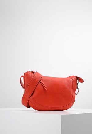 Mała czerwona skórzana torba na ramię z regulowanym paskiem, zamknięciem na zamek błyskawiczny i srebrnym okuciem, prezentowana na białym cokole.