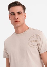 Bežové bavlněné tričko s kulatým výstřihem, krátkými rukávy a kulatým potiskem loga na levé hrudi. Hladká textura a jednoduchý design.