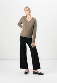Femeie îmbrăcată cu un pulover maro cu decolteu în V, pantaloni negri largi și balerini negri, stând cu o mână pe șold.