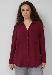 Bordeaux lange mouwen blouse met knopen van een gestructureerde stof, met een kraag en twee voorkruiszakken. Afgeronde zoom.