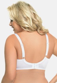 Blonde vrouw van achteren te zien, gekleed in een witte beha met verstelbare bandjes en een haak-en-oog sluiting aan de achterkant, lachend en naar links kijkend.