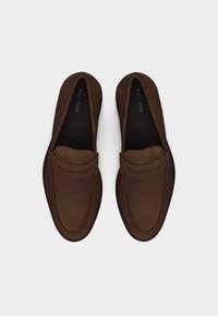 Ζευγάρι από καφέ μοκασίνια από καστόρι με μαύρες εσωτερικές σόλες με την επιγραφή "PER. ONE" σε λευκό φόντο, φωτογραφημένα από πάνω.