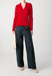 Roter, gerippter Strickpullover mit V-Ausschnitt und langen Ärmeln, kombiniert mit locker sitzenden dunklen Hosen und transparenten, belegten Sandalen. Hält eine beige Tasche.