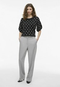 Pull en tricot noir à pois avec des manches courtes bouffantes, associé à un pantalon gris clair ajusté et des chaussures à talons noires. Fond blanc uni.