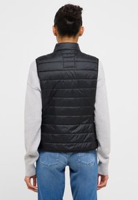 Czarna pikowana kamizelka z wysokim kołnierzem i bez rękawów, zestawiona z jasnoszarą koszulą z długim rękawem i niebieskimi dżinsami.
