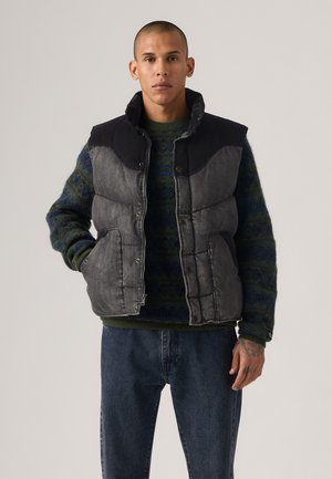 Padded gray vest with black shoulders, front snaps, and side pockets, worn over a dark green patterned sweater and blue denim pants.