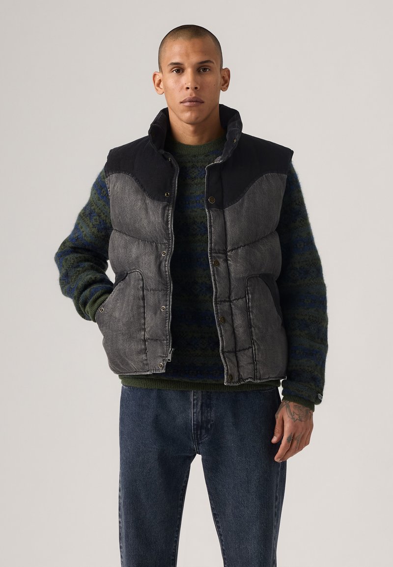 Chaleco gris acolchado con hombros negros, broches frontales y bolsillos laterales, usado sobre un suéter de patrón verde oscuro y pantalones de mezclilla azules.