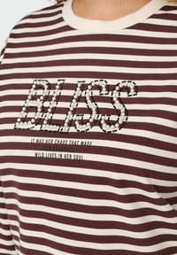 Svītrainais džemperis ir maroon un krēmkrāsā, ar teksturētu "BLISS" grafiku, kas izveidota no pērļu un kontrastējošām šuvēm.