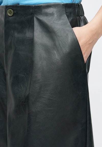 Μαύρο δερμάτινο παντελόνι με λεία υφή, διακοσμημένο με πιέτες, πλαϊνή τσέπη και κλείσιμο με πράσινο κουμπί.