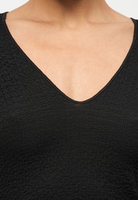 Zbliżenie na czarną, fakturowaną odzież z dekoltem w serek noszoną na gołej skórze, ukazującą górną część klatki piersiowej i okolice szyi.