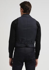 Marinblå väst med skräddarsydd passform och strukturerad design, med en spännesrem bak. Bärs över en svart långärmad skjorta.