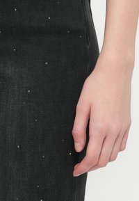 Tejido de mezclilla negra con un diseño ajustado, que presenta pequeños adornos de plata esparcidos por la superficie; mano descansando sobre el tejido.